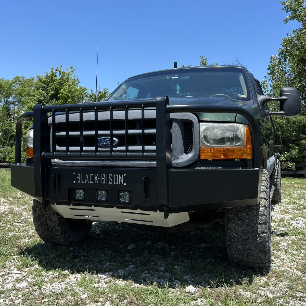 A green Ford F250 f350 pickup truck with a heavy-duty black bumper and off-road tires, parked outdoors on a gravel and grassy area under a blue sky. bumper is made by Black Bison Fab