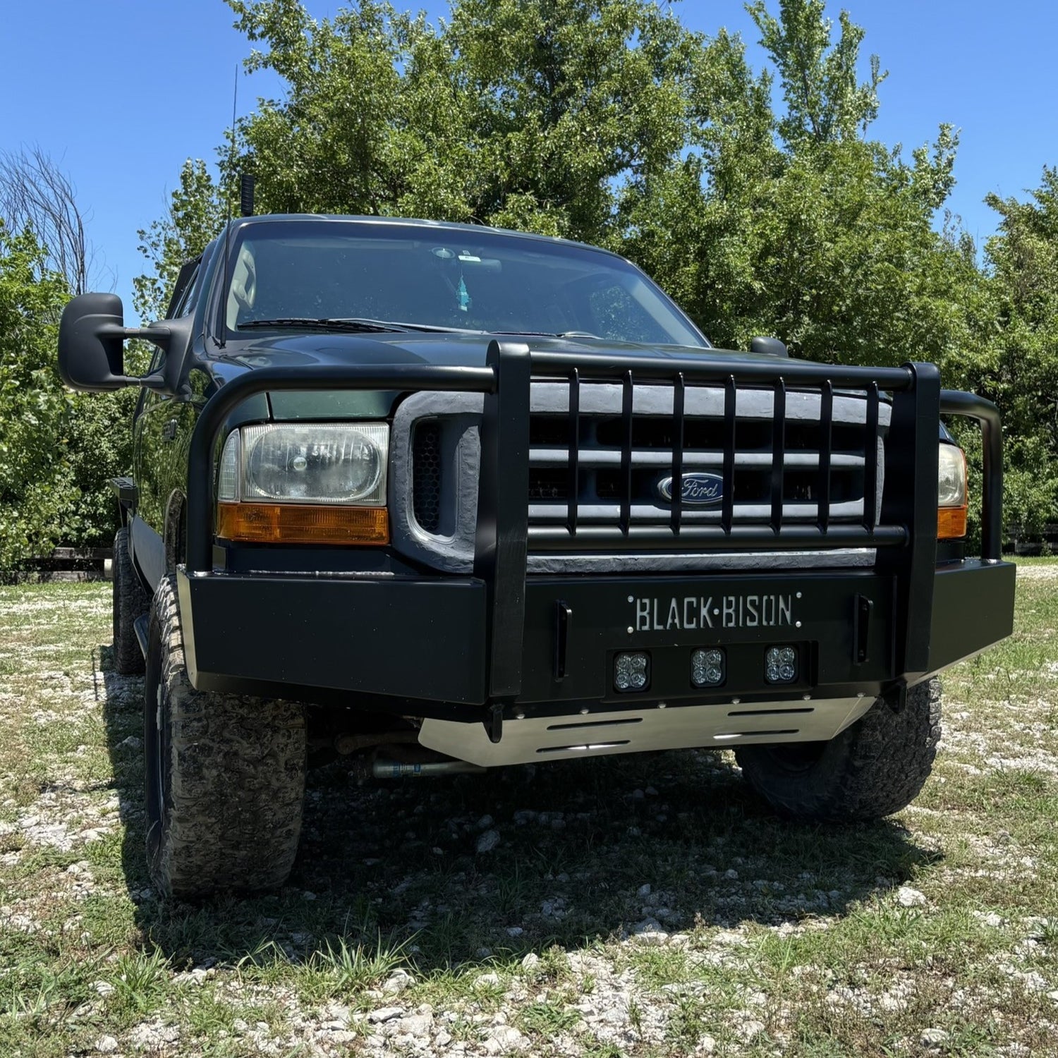 A green Ford f250 f350 pickup truck parked outdoors on grass and gravel, shown from the front angle. The truck is equipped with a heavy-duty black front bumper guard featuring the 'BLACK BISON' logo.