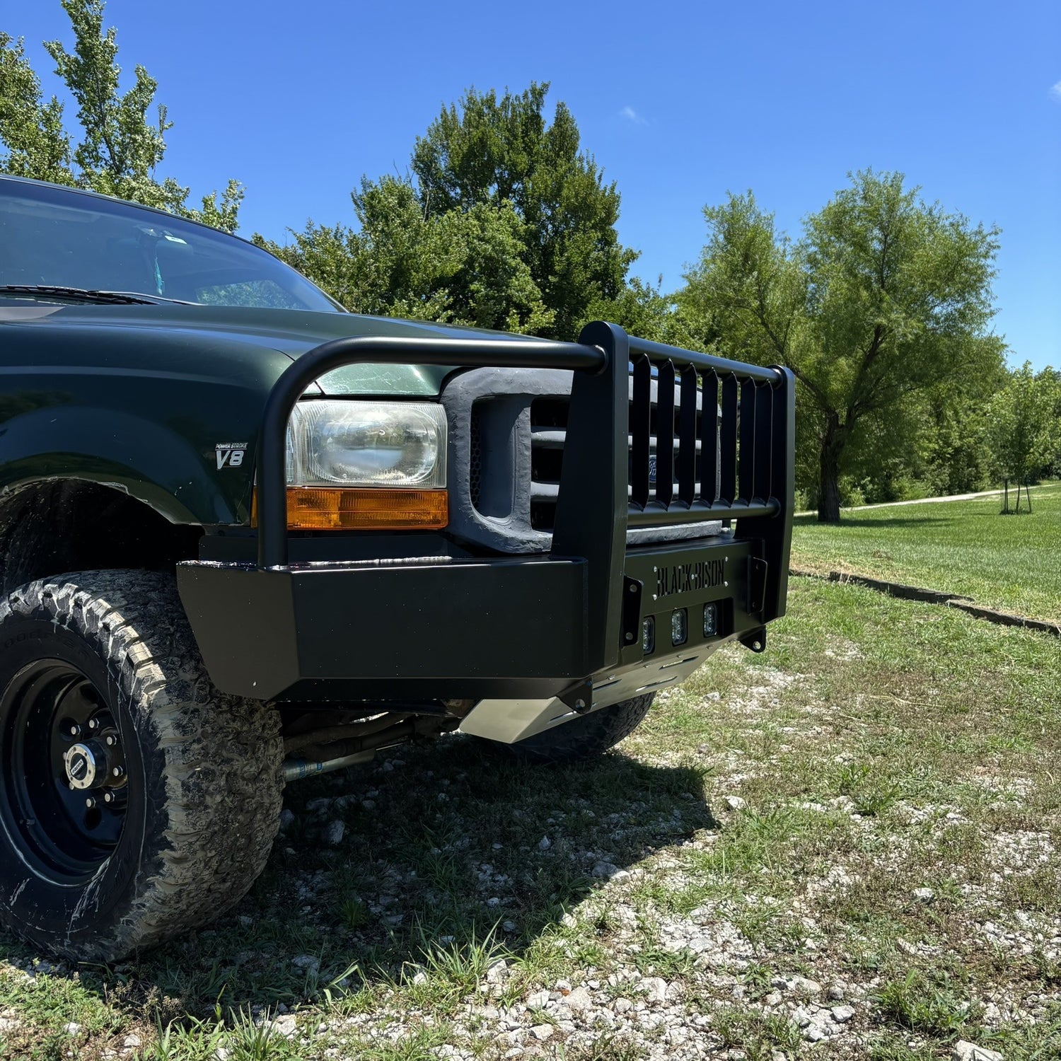 A black steel bull bar front bumper guard mounted on a dark green ford f250 f350, parked outdoors on a grassy, gravelly area with trees and blue sky in the background.