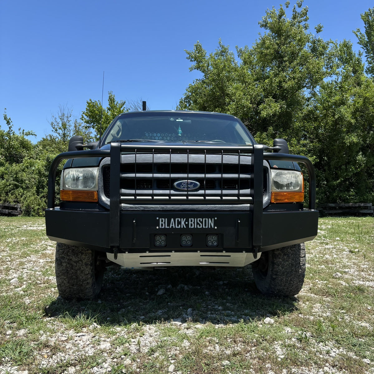 A black Ford f250 f350 pickup truck with a heavy-duty bull bar and auxiliary lights, parked outdoors on a grassy, gravelly area with trees and a blue sky in the background.