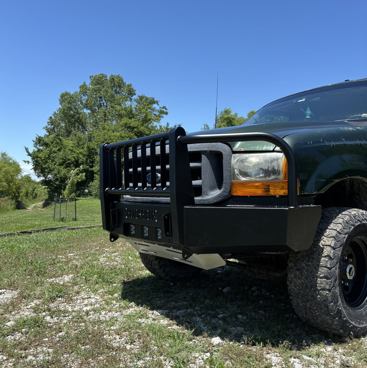 A black steel front bumper guard and bull bar installed on the front of a dark green ford f350 f250, photographed outdoors on a grassy, rocky area under a clear blue sky.