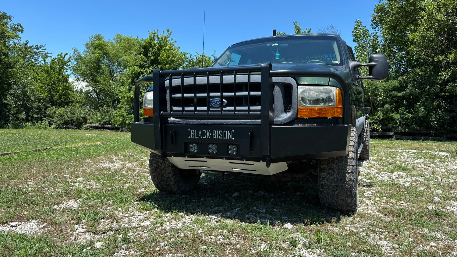 A green Ford f250 f350 pickup truck with a heavy-duty front bumper guard and off-road tires, parked on a grassy, rocky outdoor area with trees and a blue sky in the background.